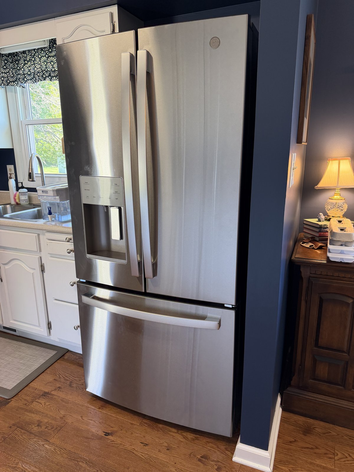 Stainless steel French-door refrigerator in a kitchen with white cabinetry, dark blue walls, and wood flooring.