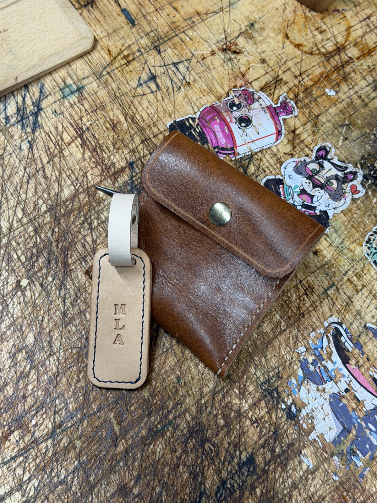 Brown leather wallet with metal snap closure and luggage tag labeled "MLA" on distressed wooden surface surrounded by colorful stickers.