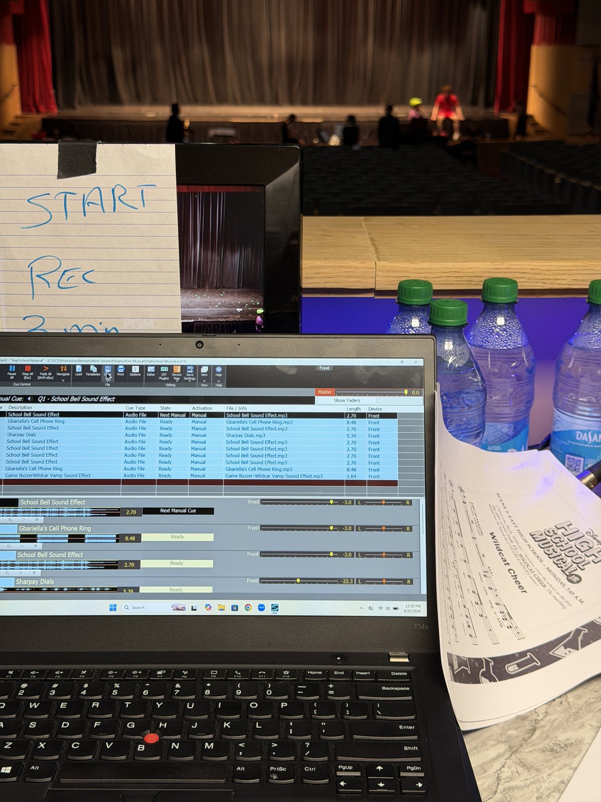 Laptop with audio editing software open showing school bell sound effects, surrounded by water bottles, sheet music, and a handwritten note on a stage in the background.