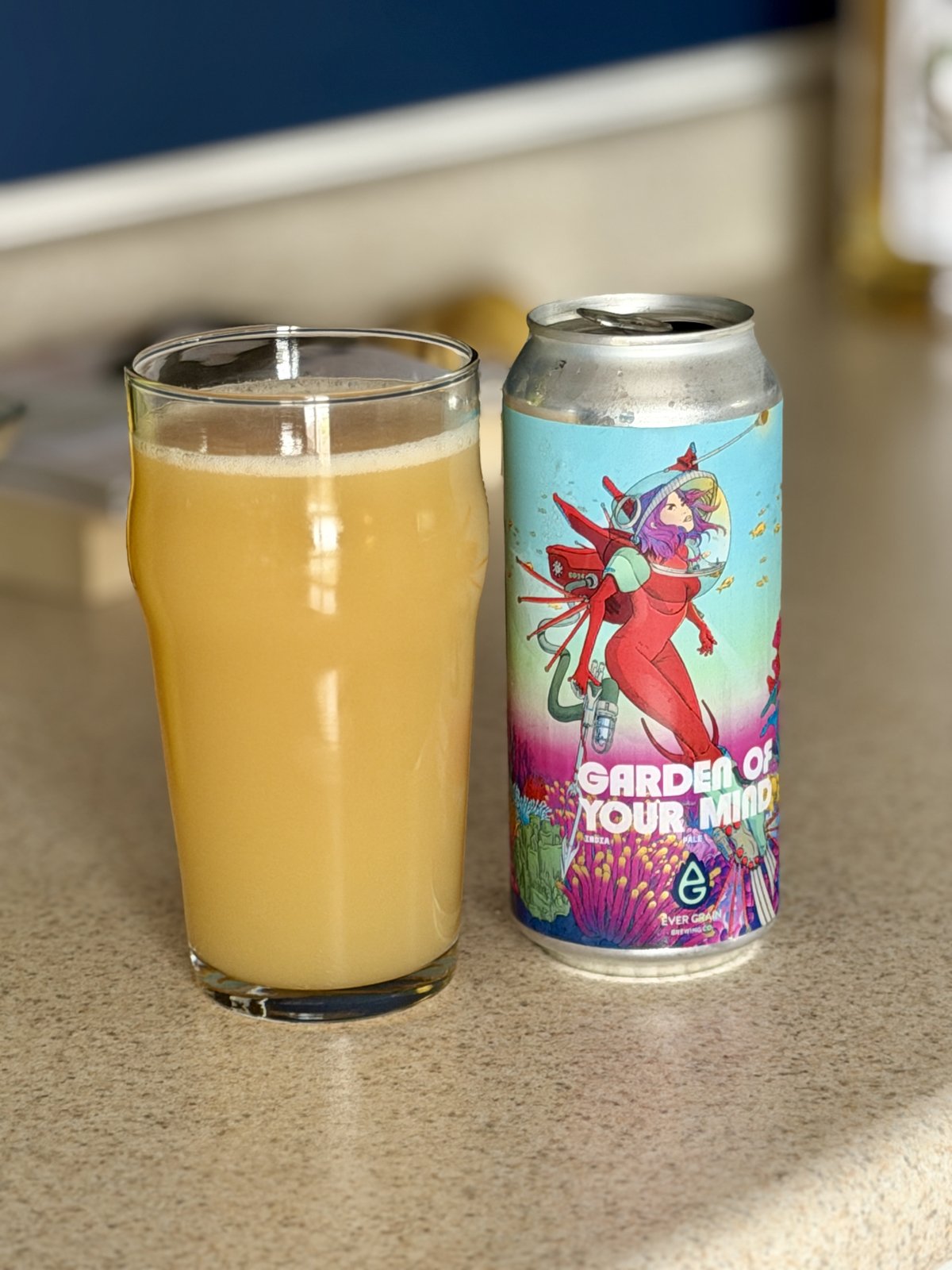 A glass of pale yellow beer sits next to a colorful craft beer can labeled "Garden of Your Mind" featuring illustrated artwork of a character in red.