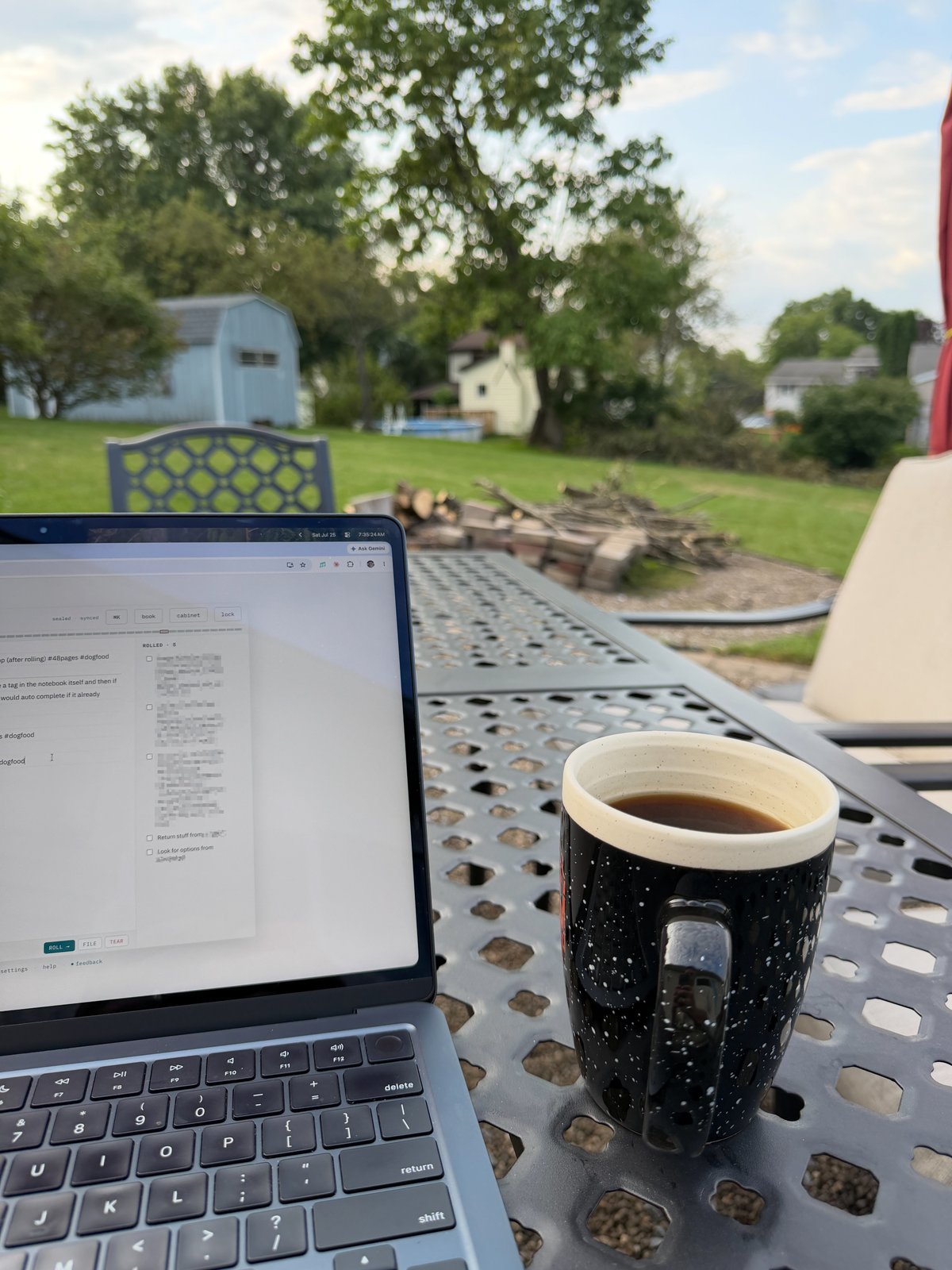 Laptop and coffee mug sit on a metal table in a backyard with trees, houses, and a fire pit visible in the background.