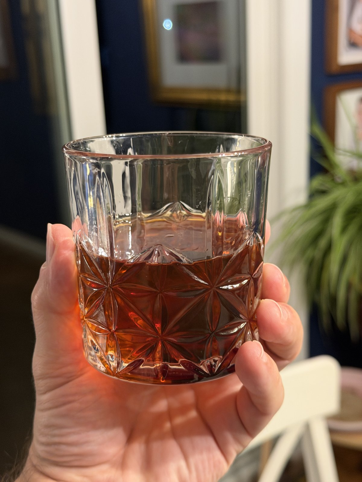 Hand holding a cut crystal glass filled with amber-colored liquor in an indoor setting with blurred background.