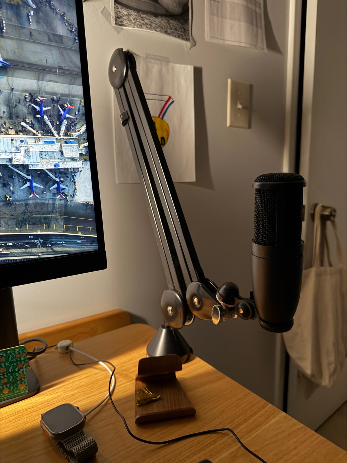 Desk setup with monitor displaying aerial view, desk arm with multiple pulleys and weights, microphone on stand, smartwatch, and cable management on wooden desk.